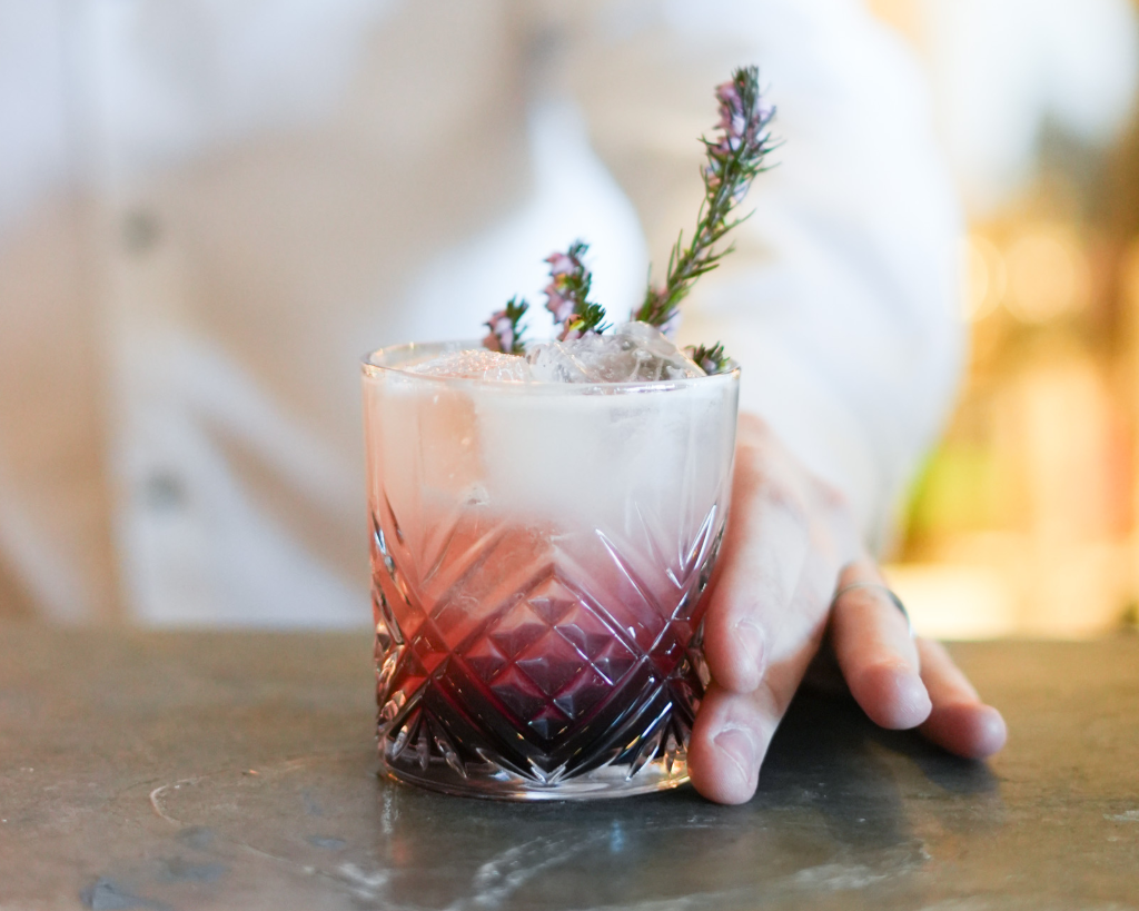 A vibrant dark purple Bramble cocktail served in a short glass on a bar counter, garnished with a lavender sprig.