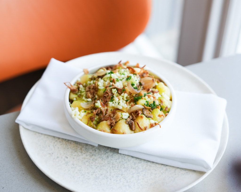 A bowl of cottage pie topped with crispy onions and crumbled cheese, served in a white dish on a folded white napkin and plate, set against a modern dining background with an orange chair. A dish served in The Brasserie.