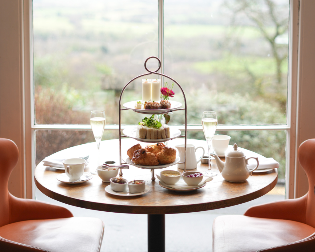 A three-tiered cake stand elegantly arranged with finger sandwiches, scones with clotted cream and jam, and an assortment of delicate cakes, accompanied by a pot of tea, and a two glasses of Prosecco on a neatly set table.