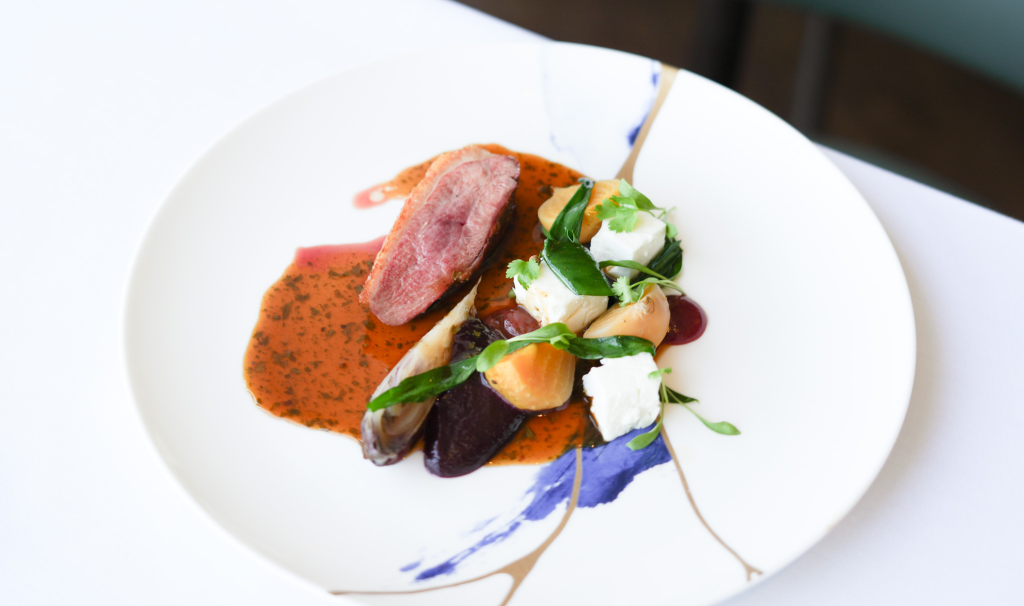 A plated fine-dining dish from 1919 Restaurant Malvern, featuring a slice of seared duck breast, accompanied by roasted vegetables, herbs, and a rich red sauce, artfully arranged on a white plate with abstract blue and gold designs.