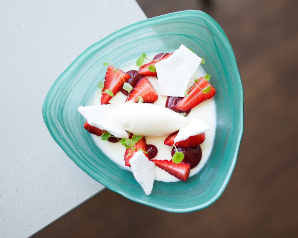A dessert from 1919 Restaurant Malvern, consisting of a white chocolate panna cotta, strawberries, vanilla ice cream and meringue pieces, served in a blue glass bowl.