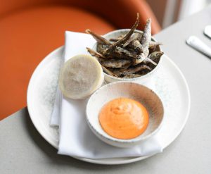 A neatly plated appetizer featuring a bowl of crispy fried whitebait, a small bowl of paprika mayonnaise, and a lemon half wrapped in mesh. The items are arranged on a folded white napkin atop a white ceramic plate, set on a light gray tabletop. In the background, there’s an orange curved chair and a window allowing natural light to illuminate the scene.