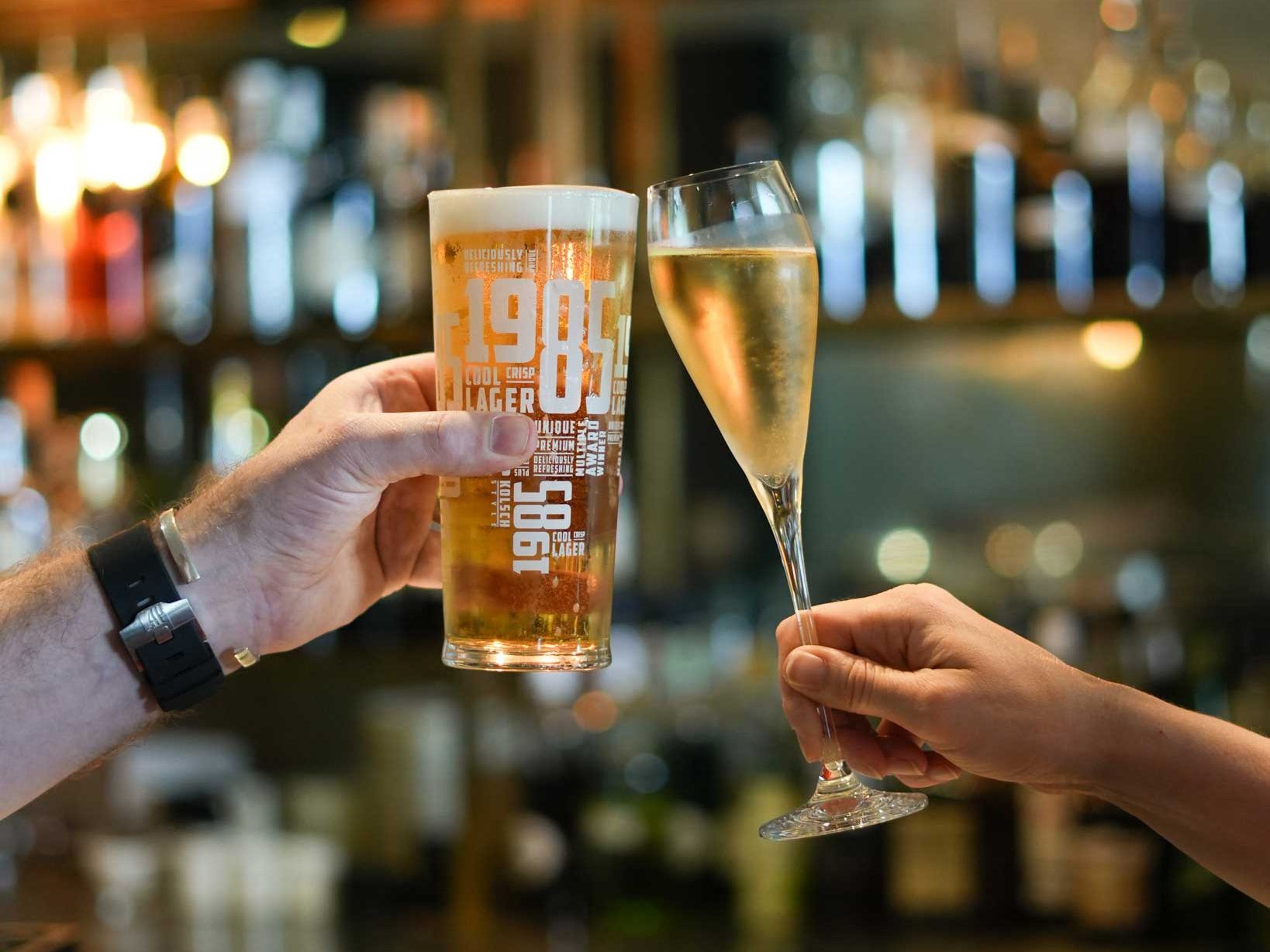 Two people clinking glasses, one with beer and the other with sparkling wine, at a bar.