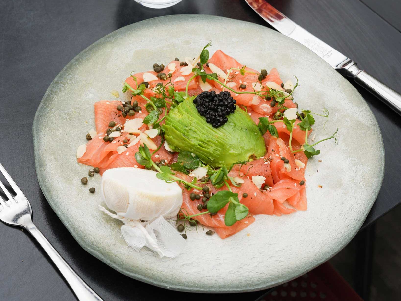 Plate of smoked salmon with avocado, caviar, capers, almonds, and microgreens, served with lemon.