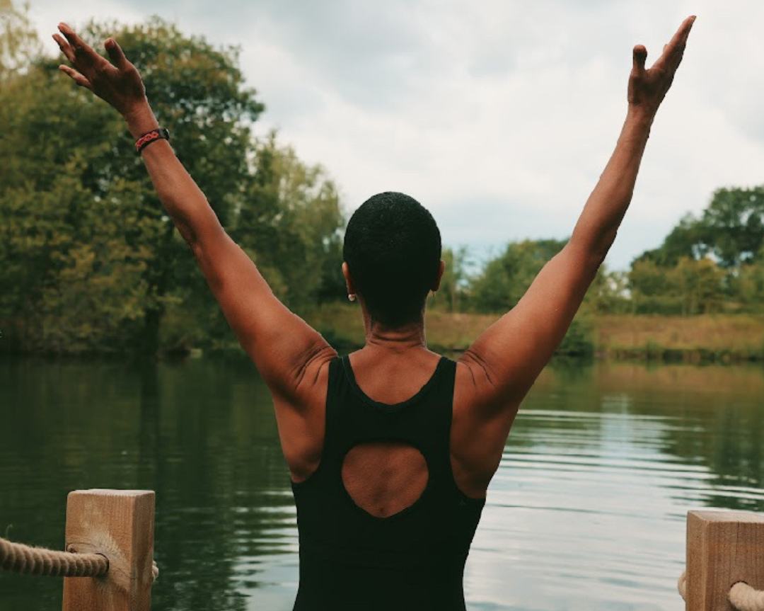 A woman standing at the edge of a calm open lake with her arms raised, preparing to start a wild swim.