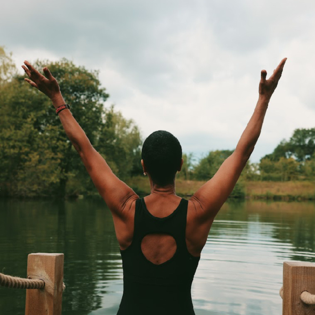 A woman standing at the edge of a calm open lake with her arms raised, preparing to start a wild swim.