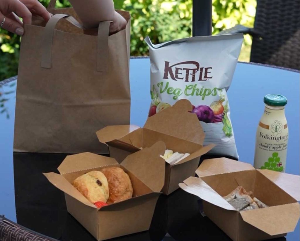 A person is reaching into a brown paper picnic bag on an outdoor glass table. On the table are open cardboard food boxes containing scones, sandwiches, and other snacks. A large bag of Kettle Veg Chips and a bottle of Folkington’s cloudy apple juice are also visible.