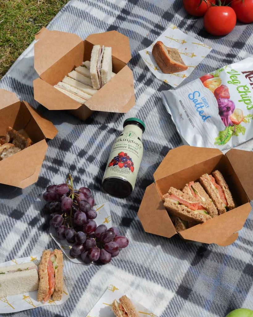 A picnic spread laid out on a blanket, featuring a selection of sandwiches, crisps, cakes, fresh fruit, and assorted drinks arranged neatly for an outdoor meal.