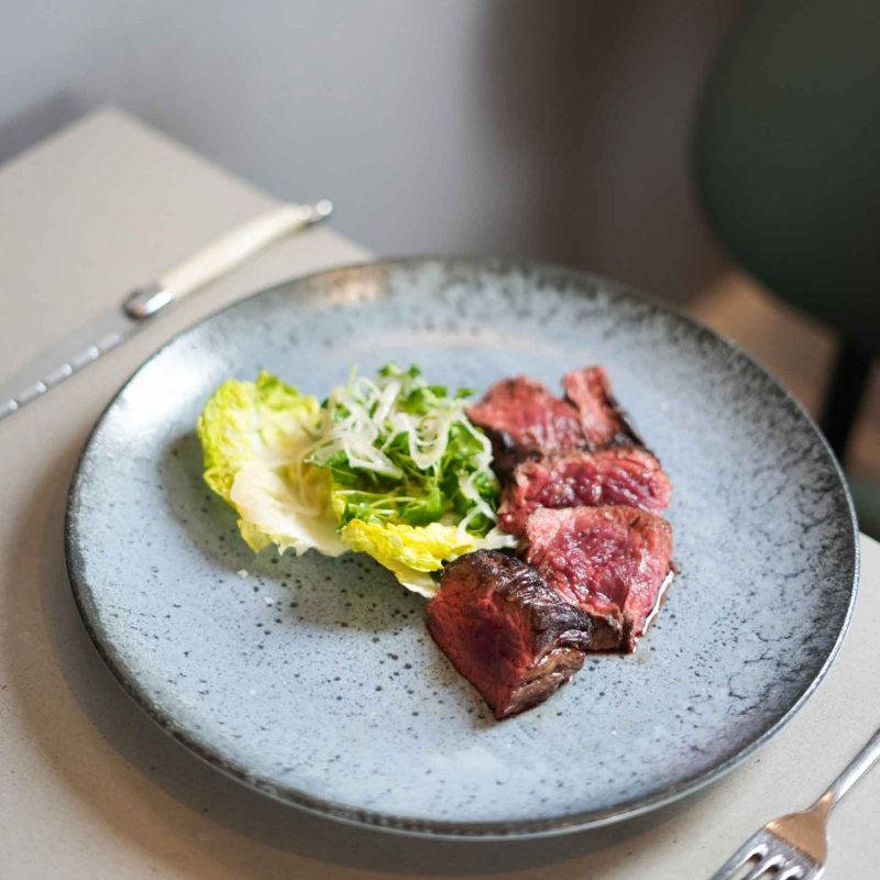 A plated, medium rare sliced steak, served with a gem lettuce salad and a chimichurri dressing. A dish served in The Brasserie.